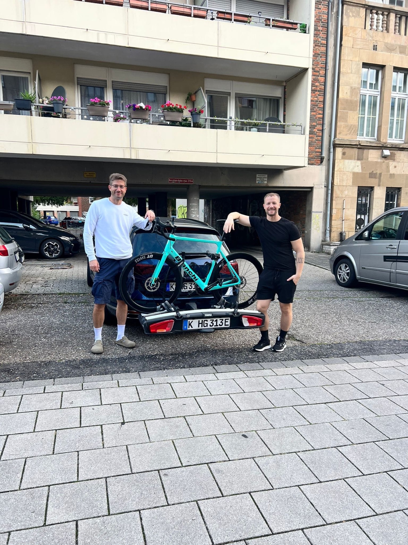 Christian und eine Begleitperson stehen neben einem Auto mit montiertem türkisfarbenem Rennrad.