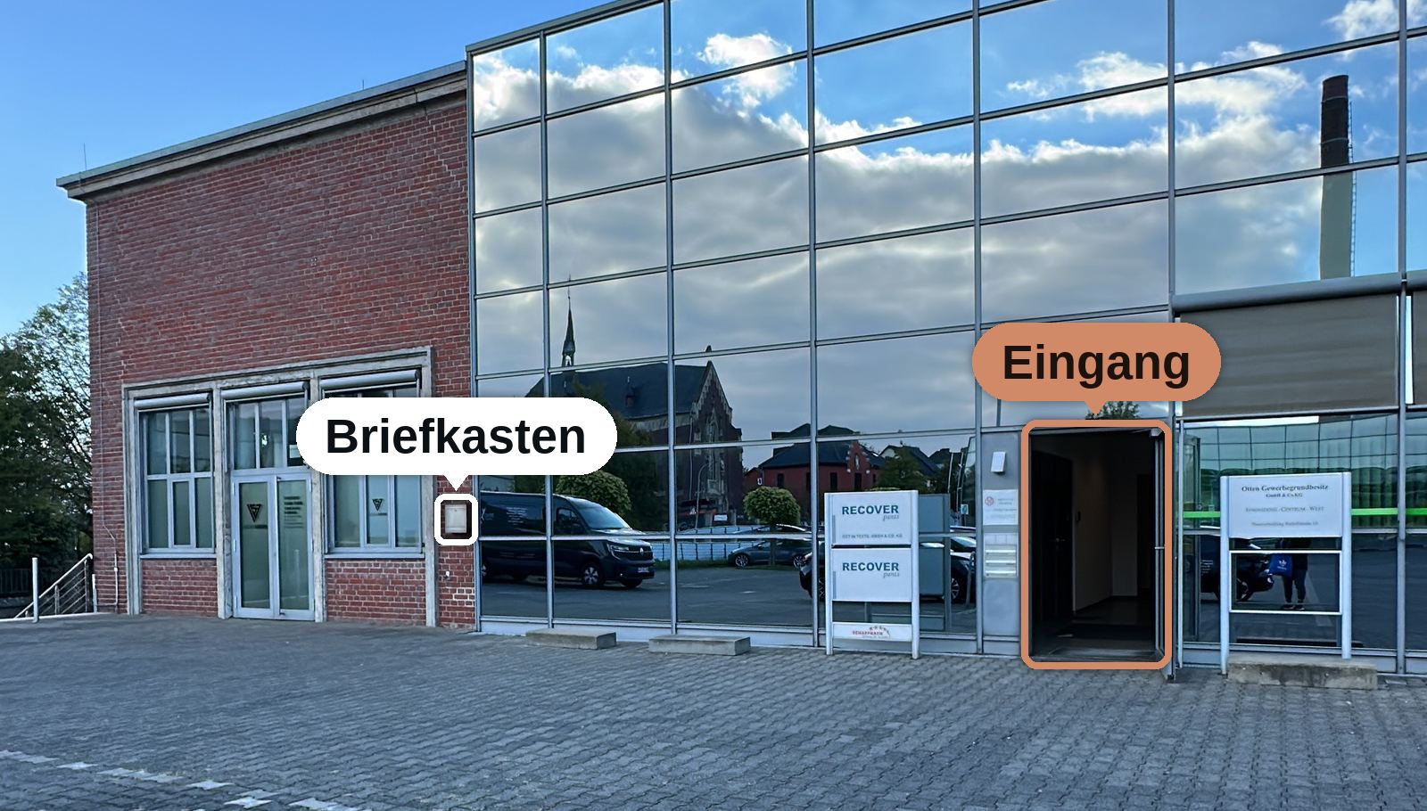 Gebäude Rudolfstraße 10C: links ein Backsteinbau, rechts ein verglaster Gebäudeteil. Die offene Glastür im verglasten Teil ist als Eingang markiert, der silberne Briefkasten am Backsteinbau neben der Glasfassade ist ebenfalls markiert.