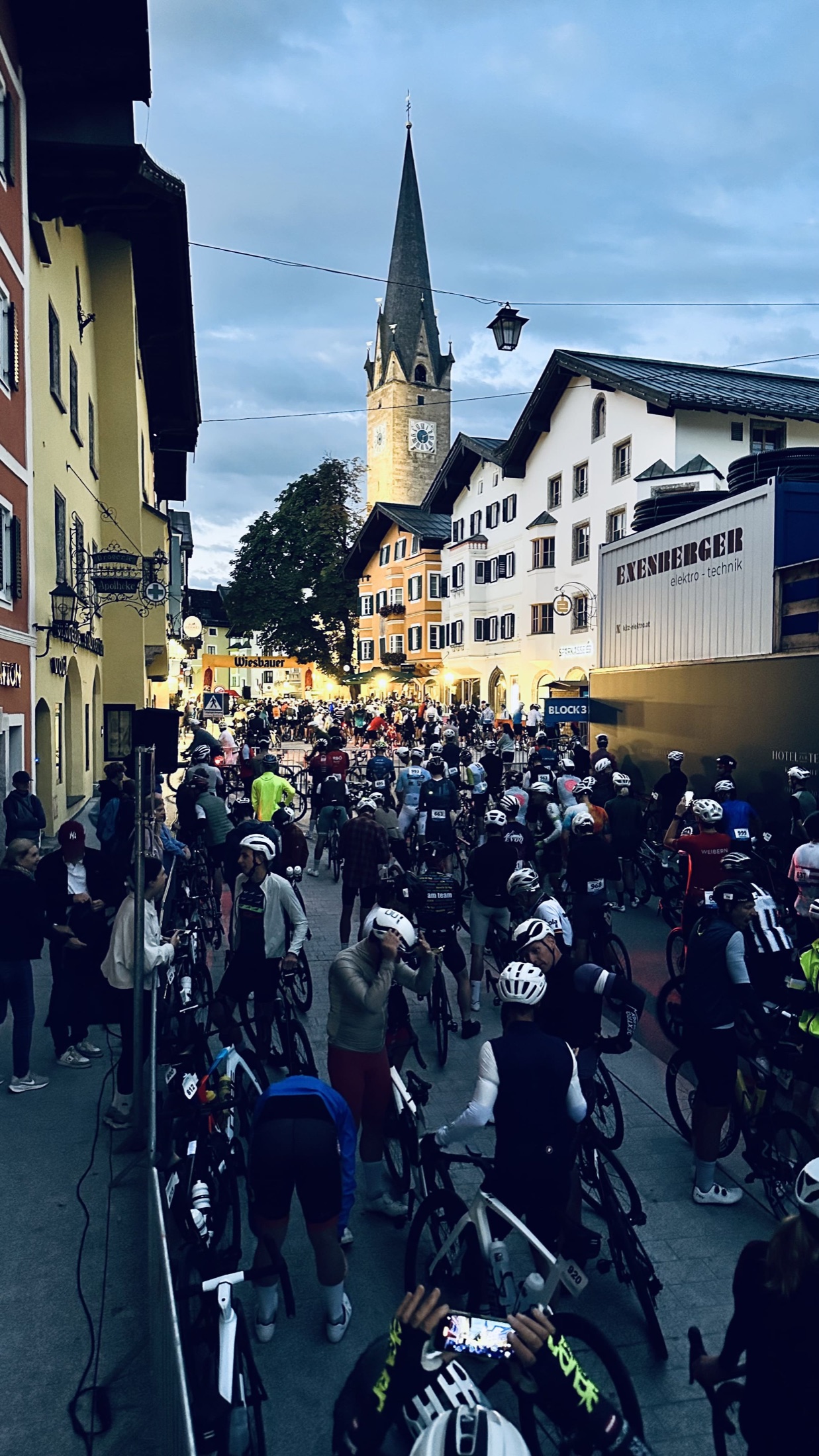 Ein großes Fahrerfeld mit Rennrädern wartet vor der beleuchteten Kirche in Kitzbühel.