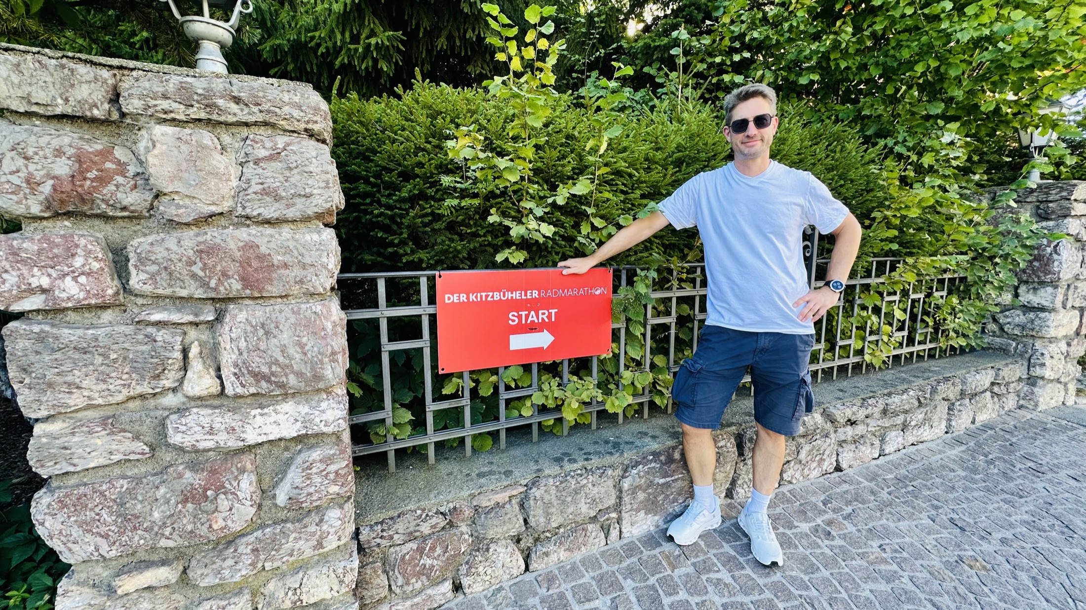 Christian steht neben einem roten Wegweiser mit der Aufschrift Der Kitzbüheler Radmarathon – Start.