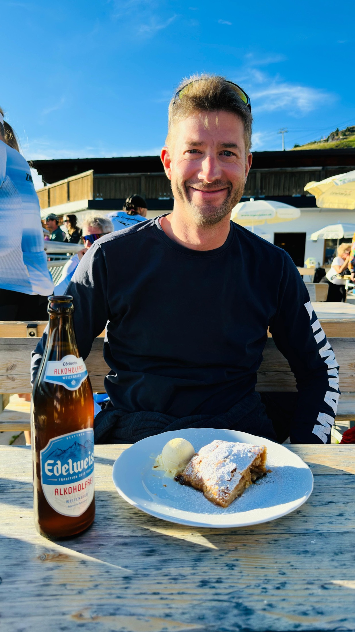 Christian sitzt lächelnd an einem Tisch vor alkoholfreiem Weizenbier und einem Teller mit Strudel und Eis.